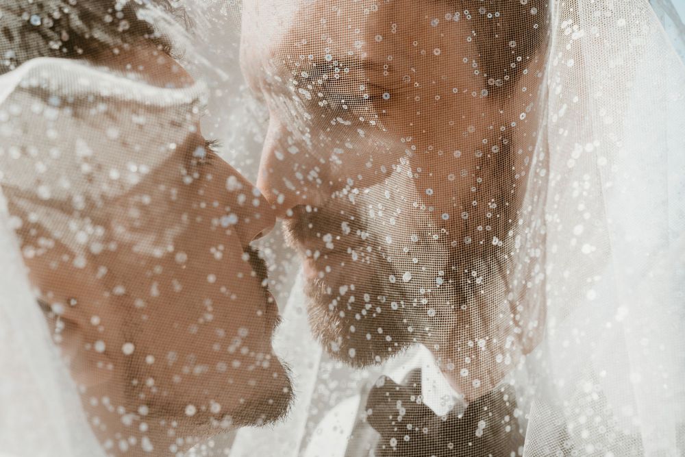 Romantic wedding couple kissing under a sheer veil with sparkling water droplets creating a dreamy bokeh effect.