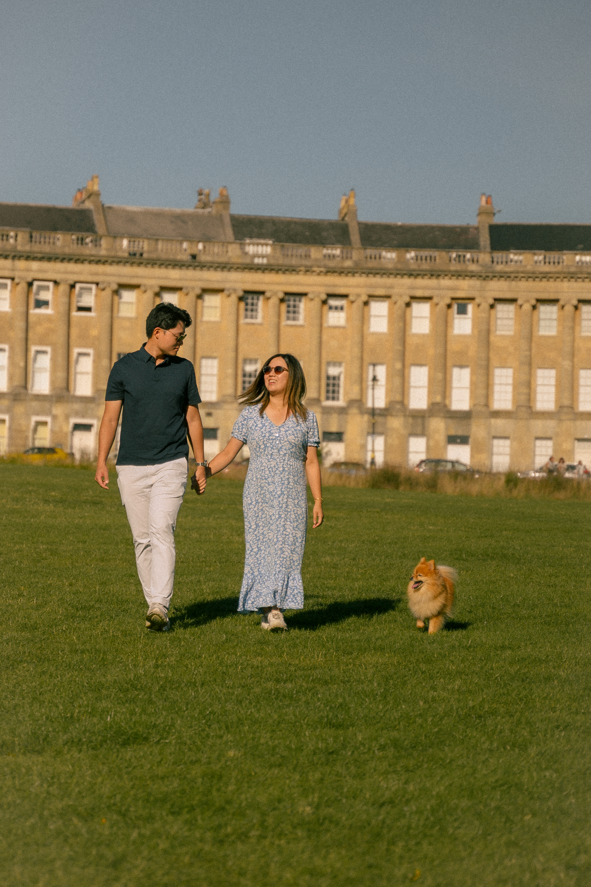 A couple holds hands walking a Pomeranian dog on green grass in front of the Royal Crescent in Bath, England.