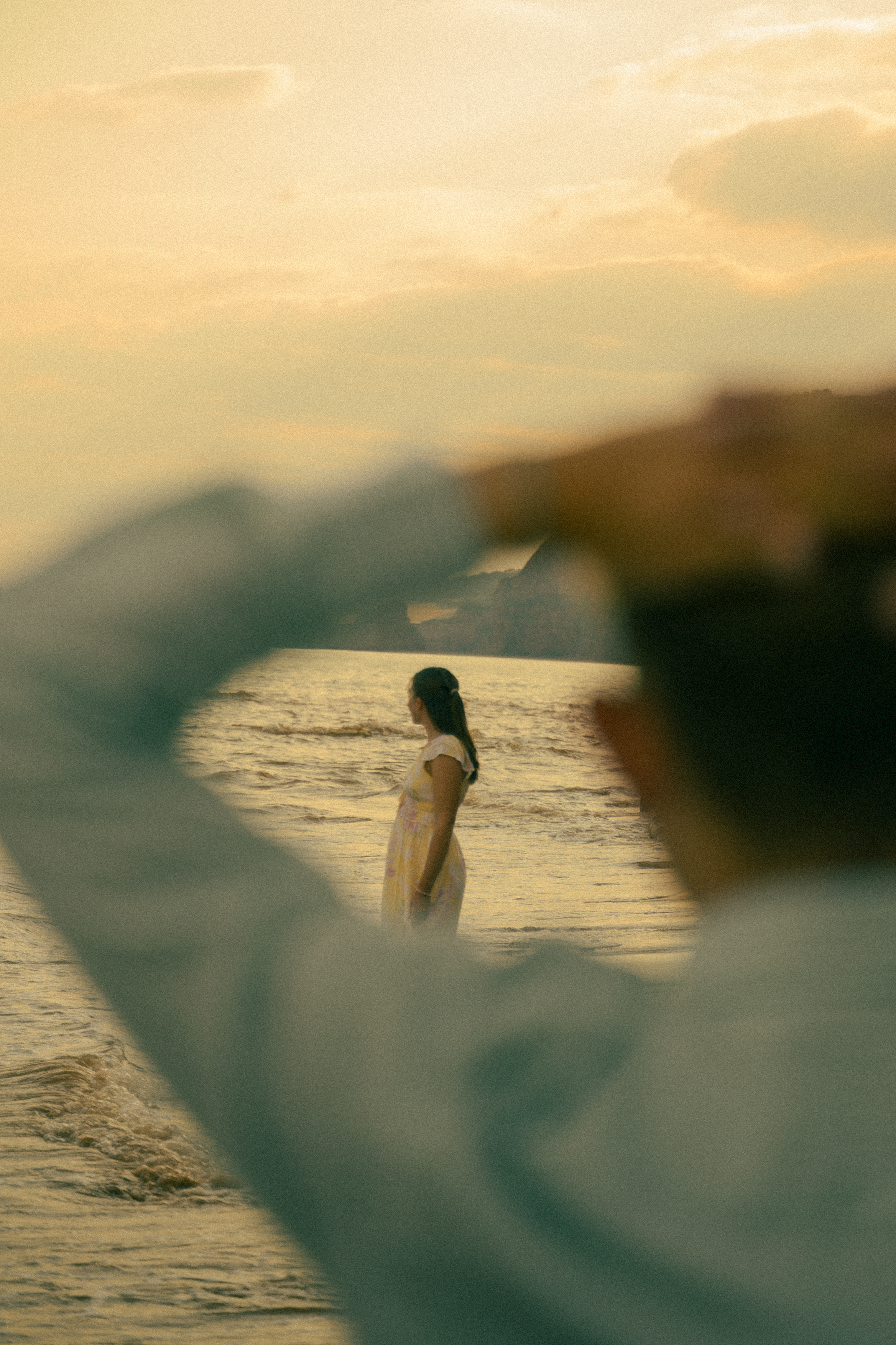 A woman in a white dress stands by the ocean at sunset, viewed through a blurred foreground frame.