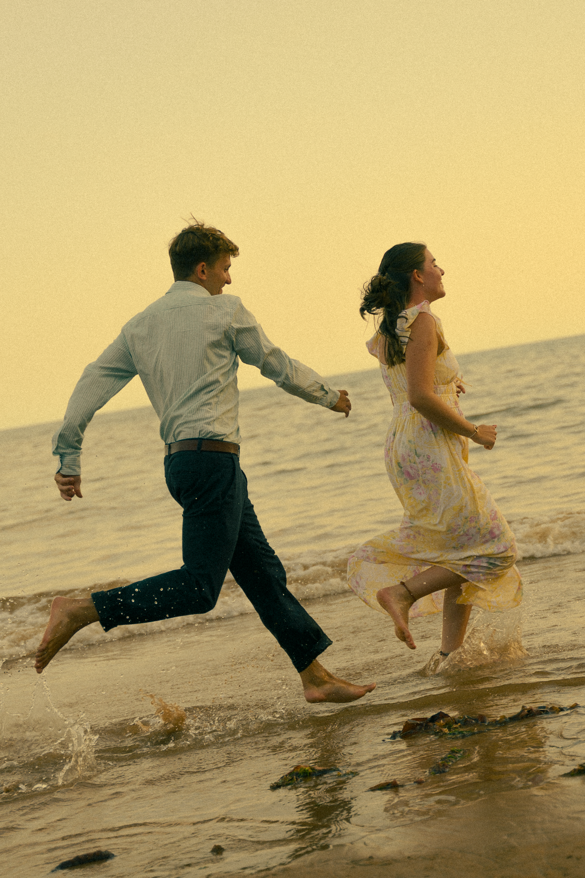 A couple runs barefoot along the ocean shoreline at sunset, holding hands in a joyful, romantic moment.
