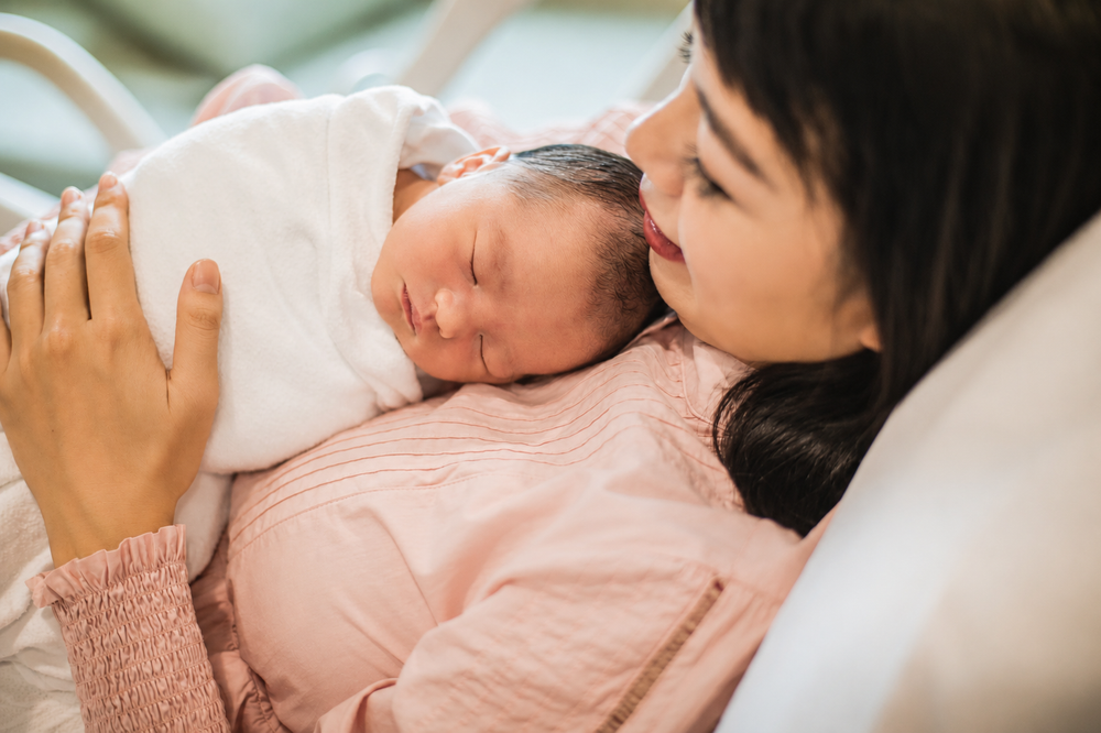 Close-up of Asian mother gazing lovingly at sleeping newborn baby resting on her chest in hospital bed.