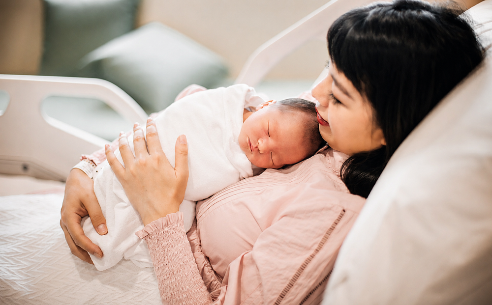 Asian new mother resting in hospital bed holding sleeping newborn baby wrapped in white blanket after delivery.