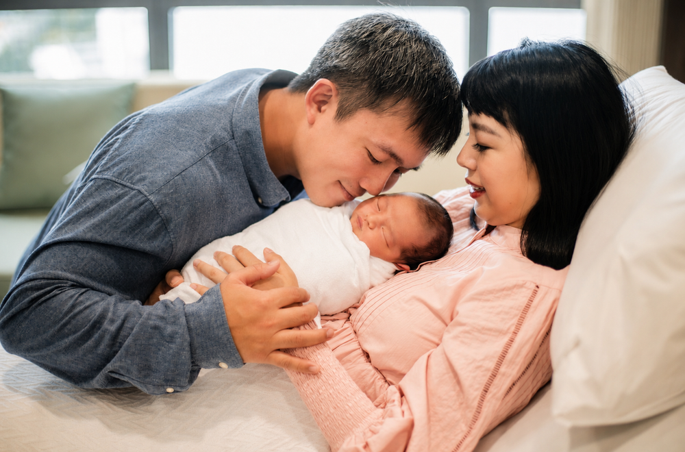 Happy Asian couple kissing their swaddled newborn baby in hospital.