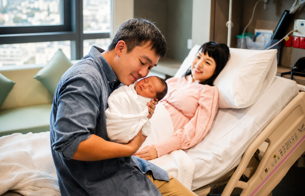 Proud Asian parents smiling at their newborn in a hospital room.