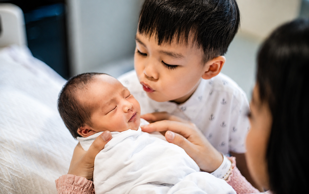 Young Asian boy gently kisses newborn sibling held by mother.