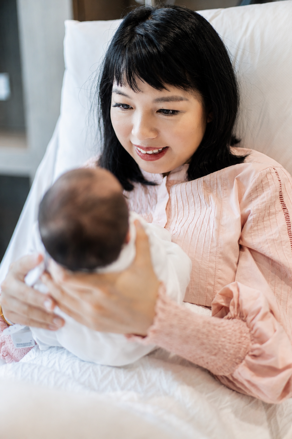 Smiling Asian mother in pink gown lovingly holds her newborn baby.