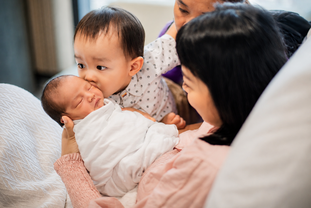 Asian mother with toddler and newborn baby bonding in hospital bed.