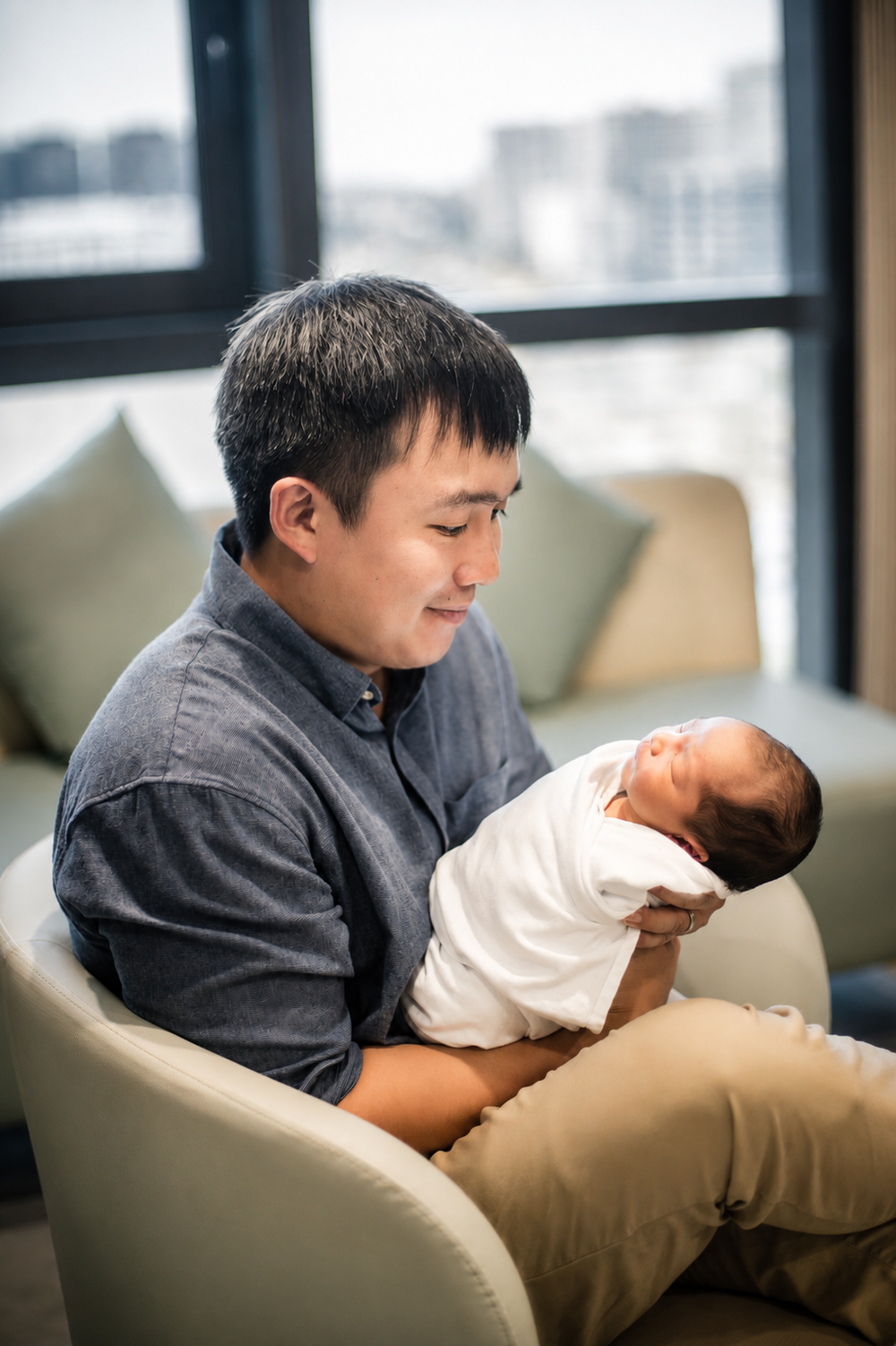 Asian father sitting in chair lovingly holding his swaddled newborn baby.