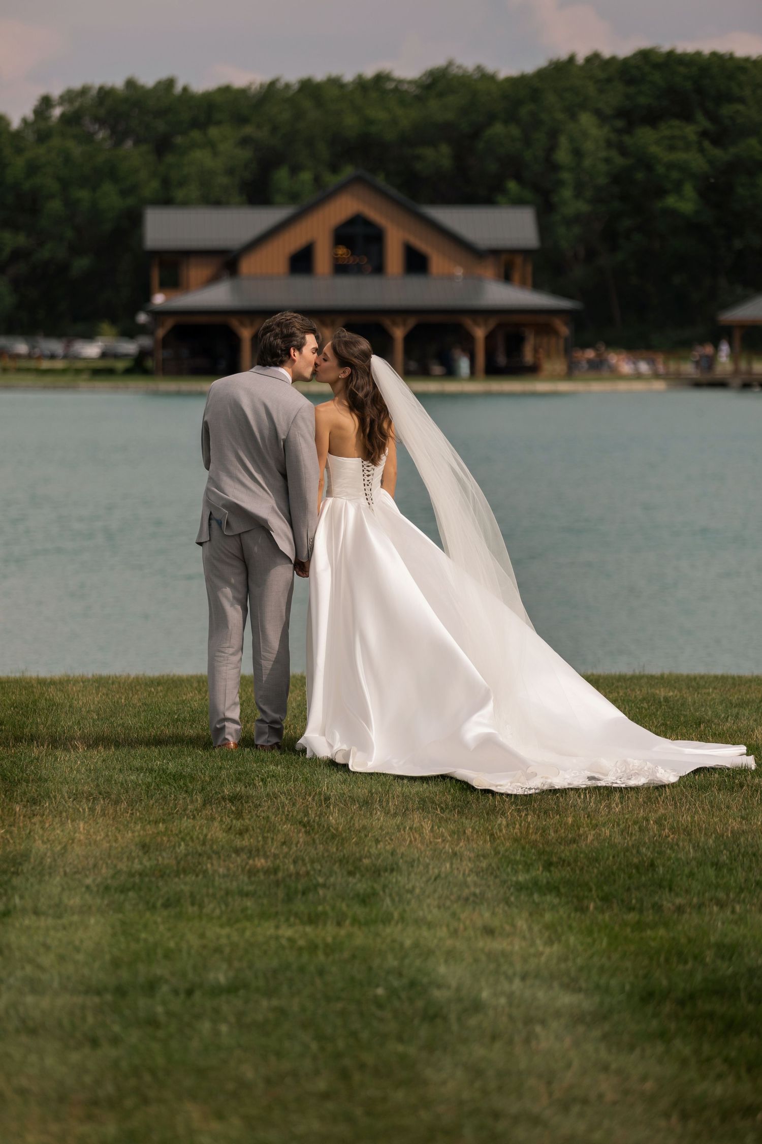 Bride and groom embracing on a wooden lakeside dock pavilion with Adirondack chairs and string lights at dusk.