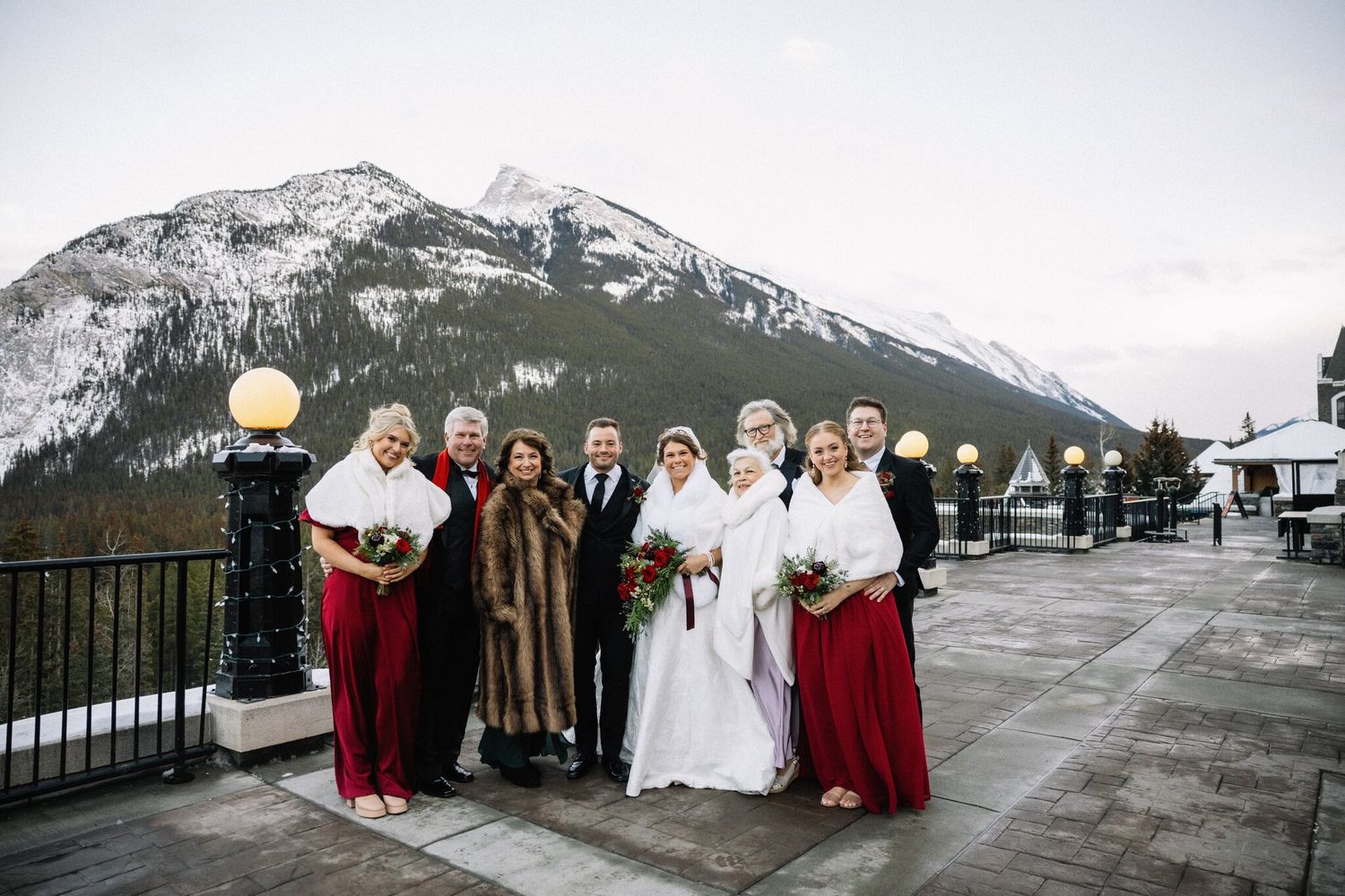 Bride and groom with family and two bridesmaids in red on a Fairmont Banff Springs terrace beneath a mountain