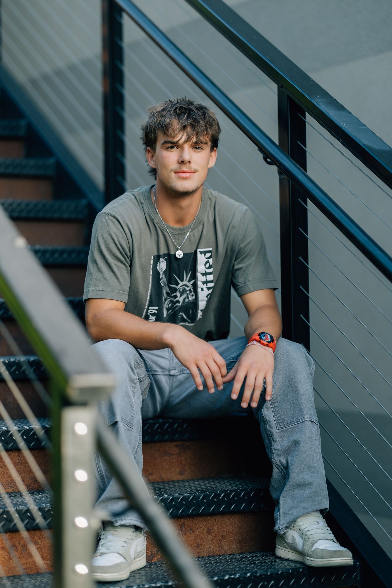 Young man in gray graphic tee and jeans sitting on metal stairs, wearing an orange watch and white sneakers.