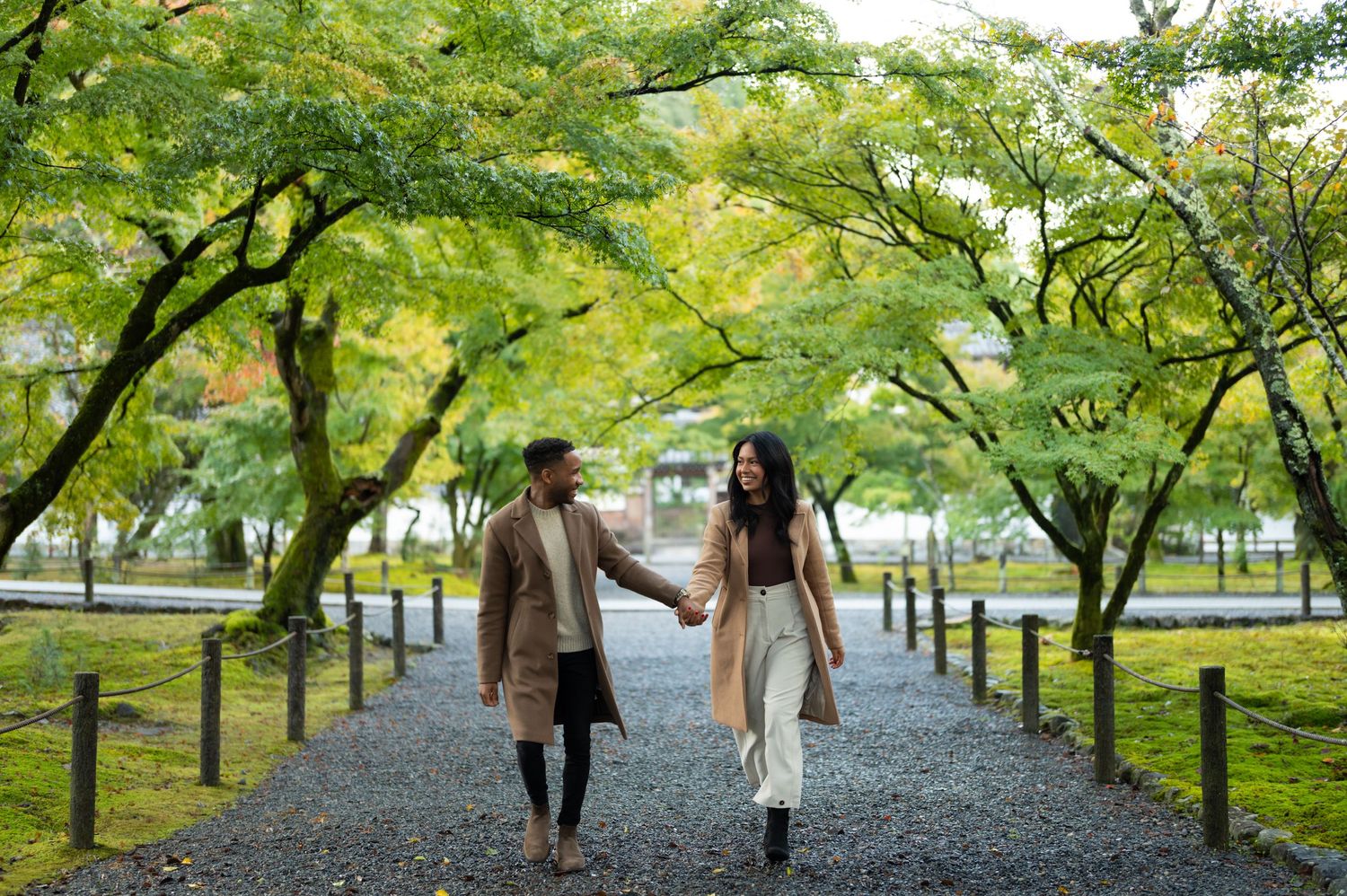 Couple holding hands walking along a tree-lined gravel path in a lush Japanese garden during autumn.