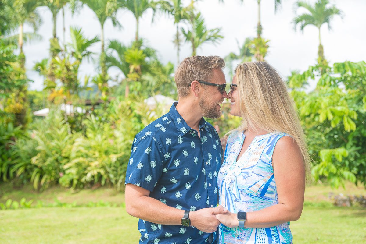 Smiling couple in matching blue tropical outfits share an affectionate moment among lush palm trees and green tropical gardens.
