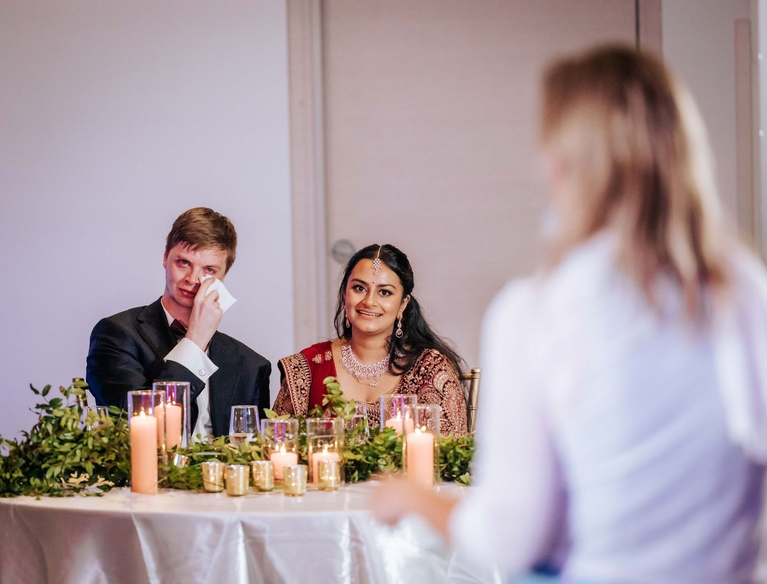 Groom dabs his eyes with a tissue beside his smiling bride at a candlelit head table while a speaker addresses them at a wedding reception.