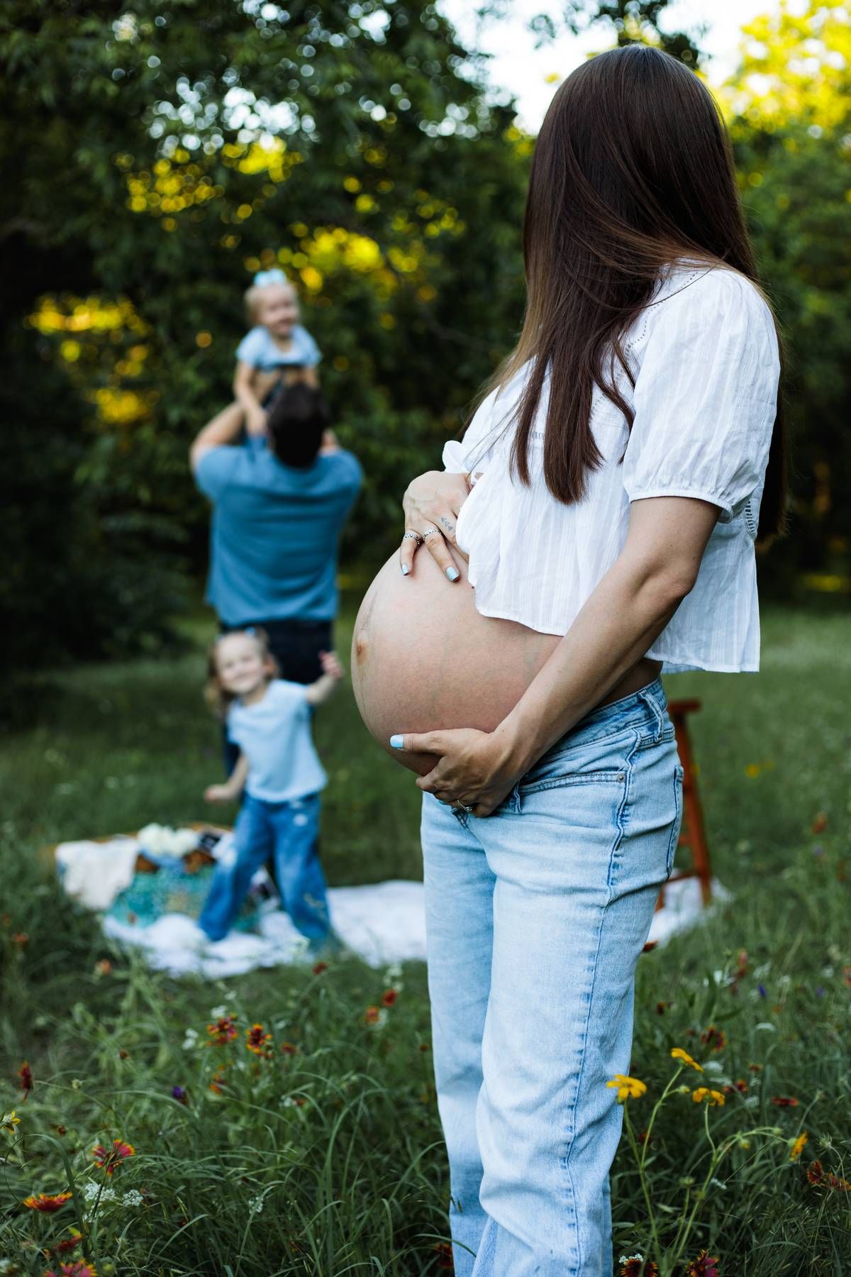 Pregnant mother cradles her belly in the foreground while a father lifts a toddler and another child plays on a meadow blanket.