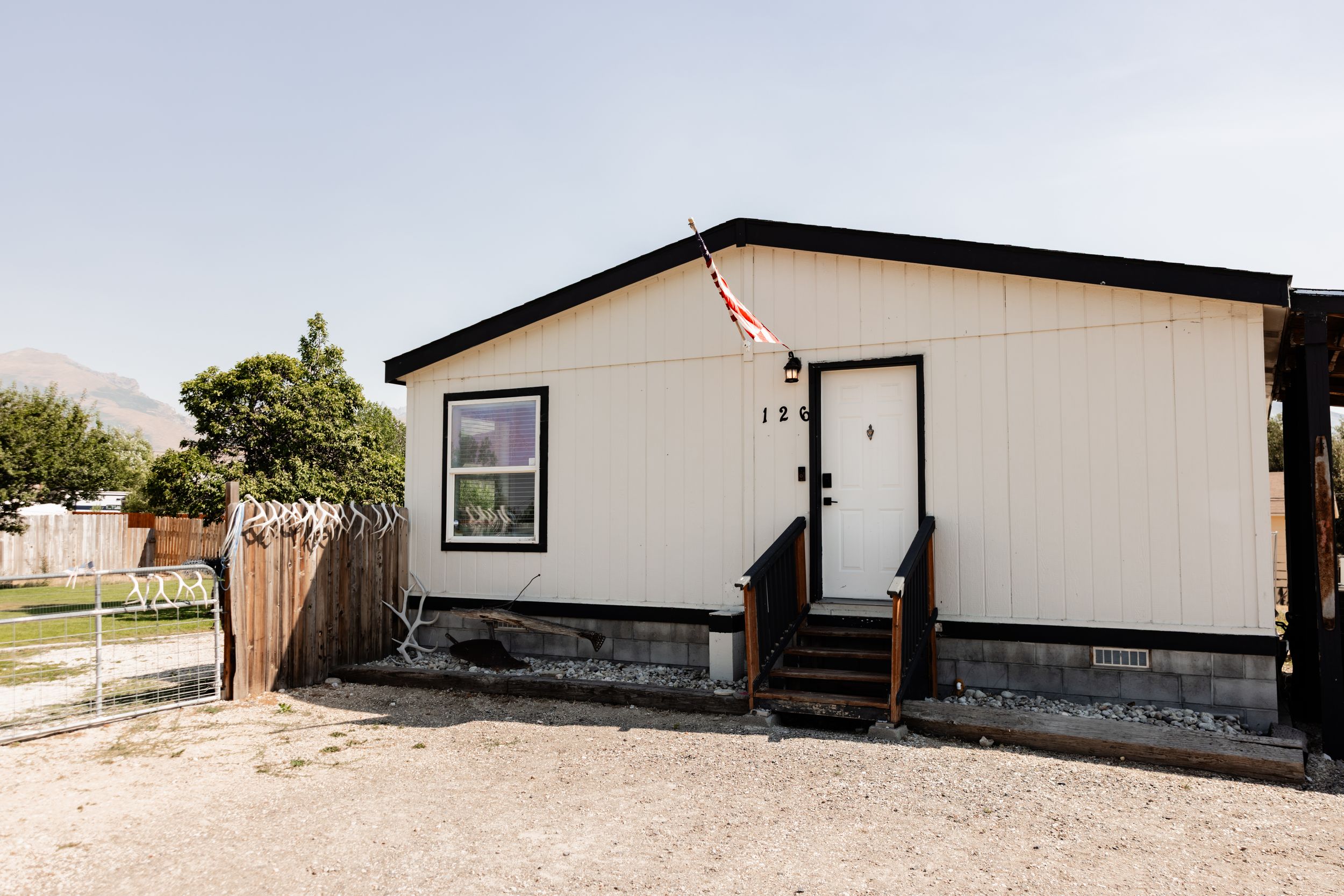 Single-story manufactured home with cream siding, black trim, wooden front steps, and a fenced yard with mountain views in the background.