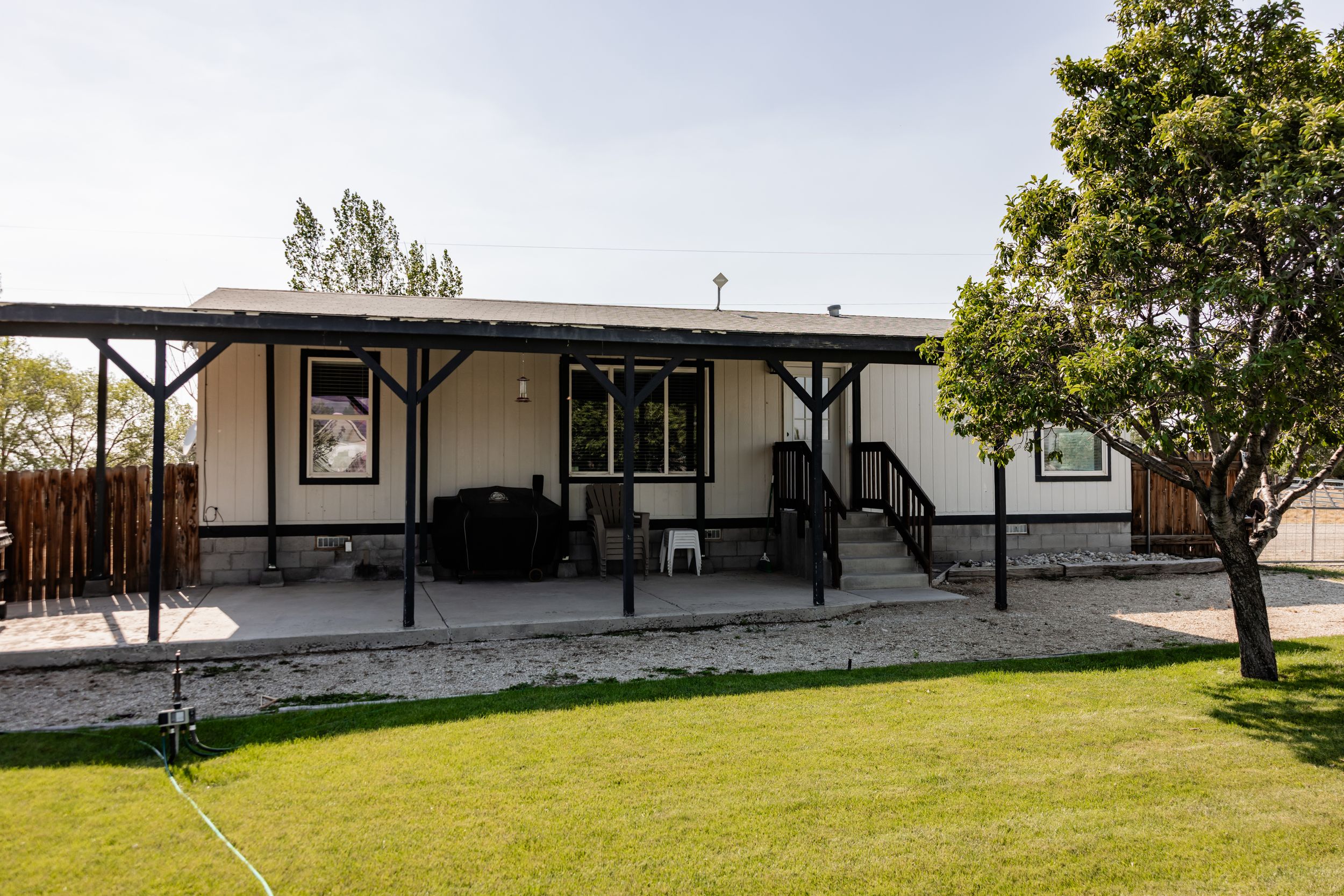 Single-story manufactured home with a covered back porch, patio furniture, wooden steps, and a green lawn beside a mature shade tree.
