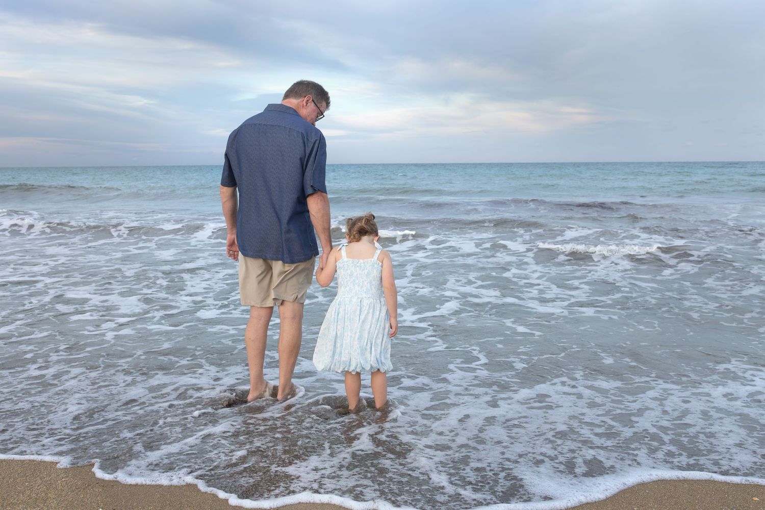 A man in a blue shirt and shorts holds hands with a little girl in a white dress as they stand in the shallow ocean surf.