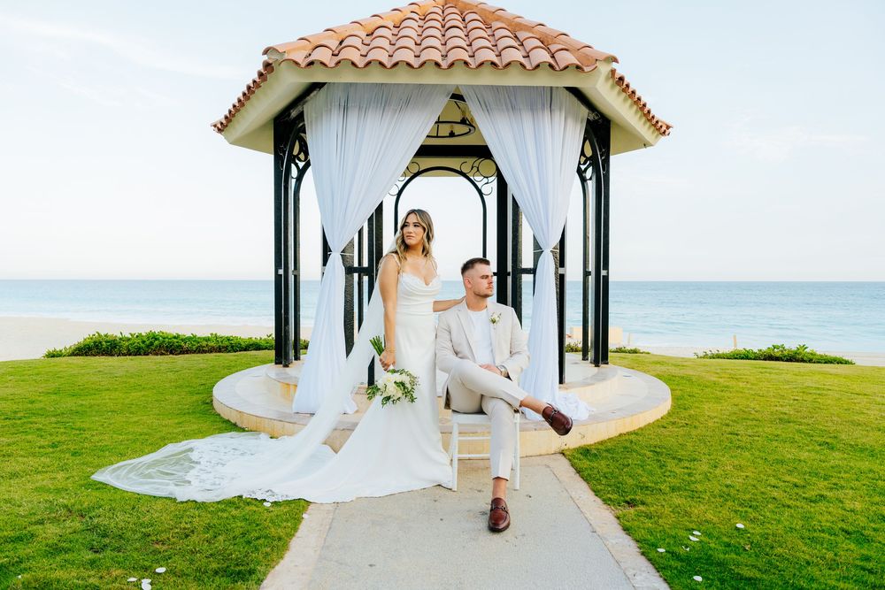 Bride in a flowing white gown stands behind a seated groom in a light suit at a beachfront gazebo with draped curtains and ocean views.
