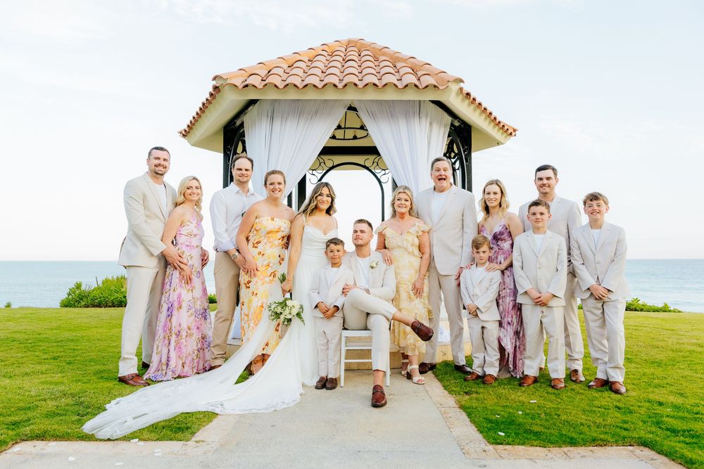 Bride and groom seated on gazebo steps surrounded by smiling family in pastel and neutral attire at an oceanfront beach wedding.