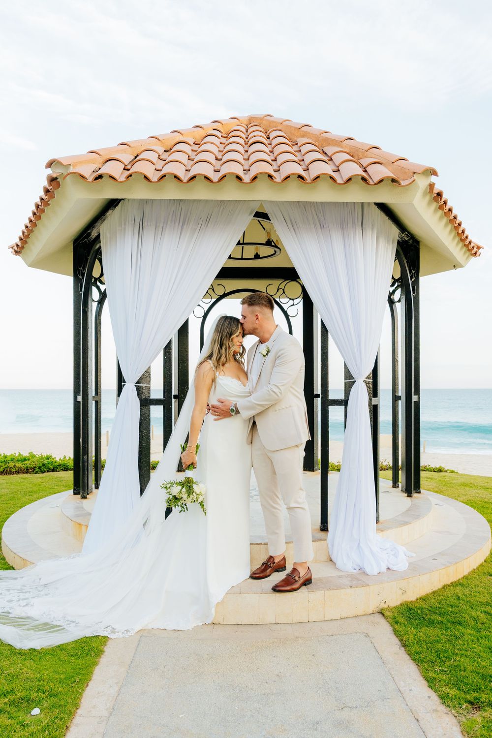 Bride and groom embrace forehead to forehead inside a white-draped beachfront gazebo with a terracotta roof overlooking the ocean.