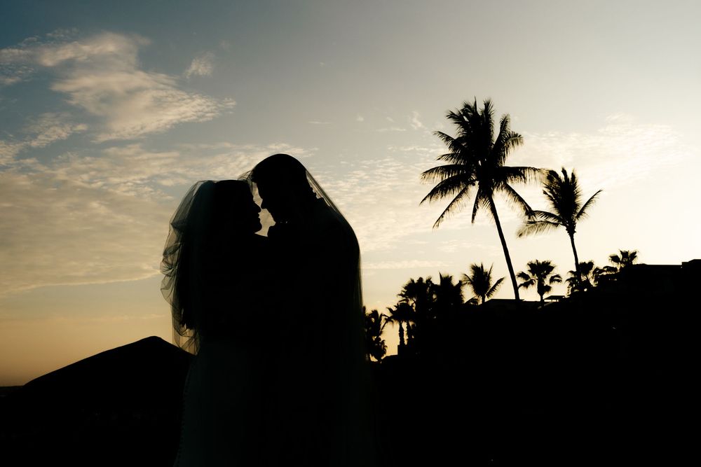 Silhouette of a bride and groom embracing at sunset with palm trees and a glowing orange sky in the background.