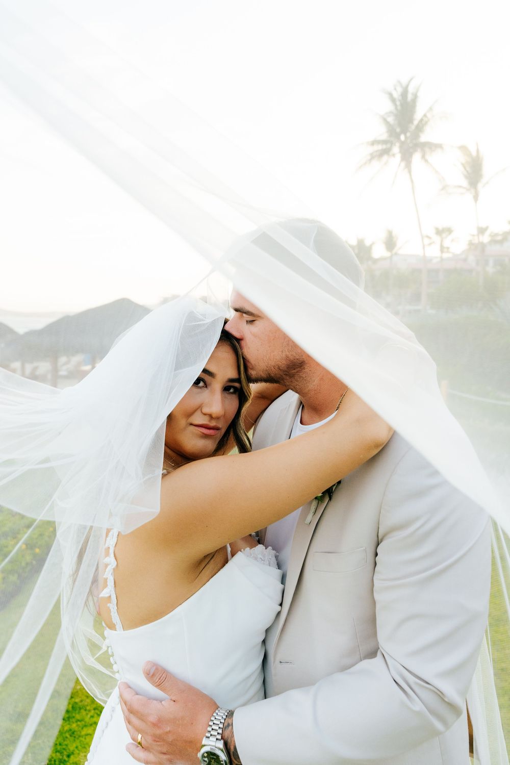 Bride and groom embrace under a sheer wedding veil as he kisses her forehead, with palm trees and a tropical resort behind them.