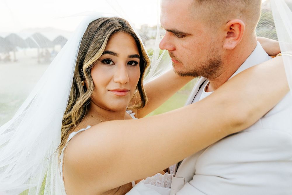 Bride in a white veil and groom in a light suit embrace closely in a romantic wedding portrait with a blurred tropical backdrop.