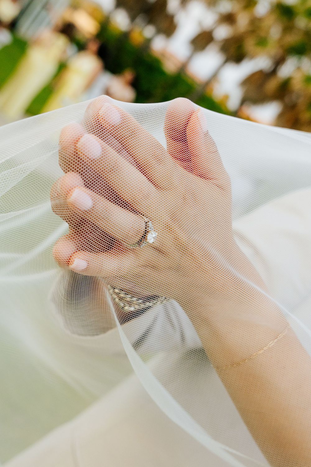 Close-up of a bride's hands with pale pink manicure and a diamond engagement ring, softly draped in a sheer wedding veil.