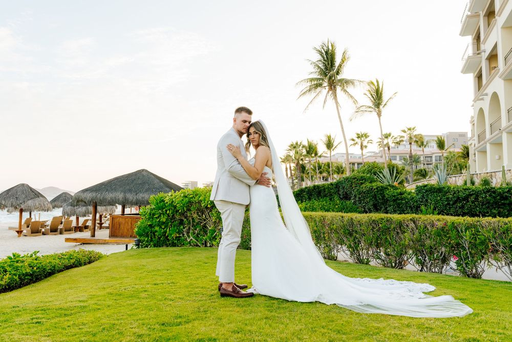 Bride and groom embrace on a lush green lawn at a tropical beach resort wedding, with palm trees and thatched palapas behind them.