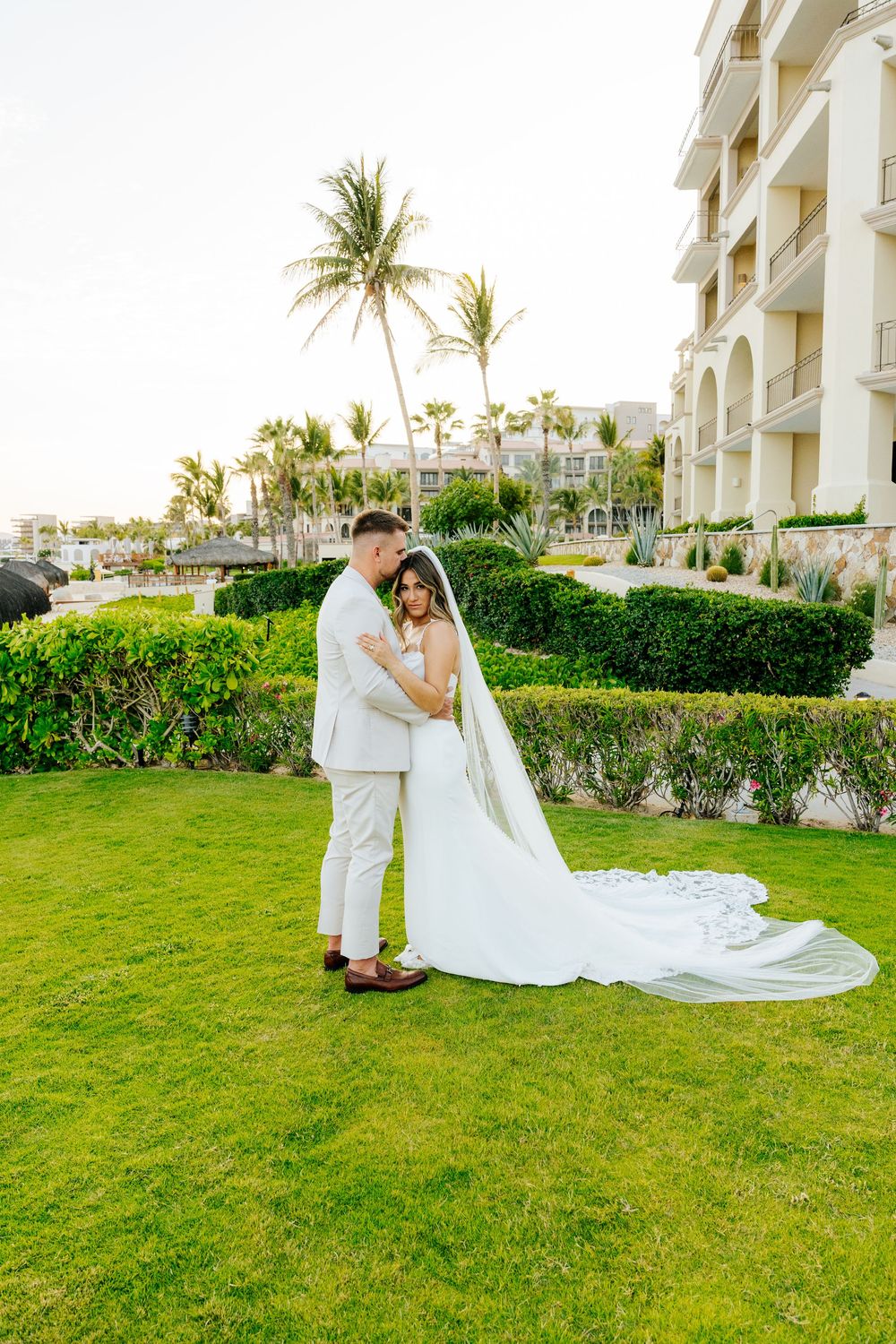 Bride in a flowing white gown and long veil embraces her groom in a cream suit on a lush resort lawn with palm trees.