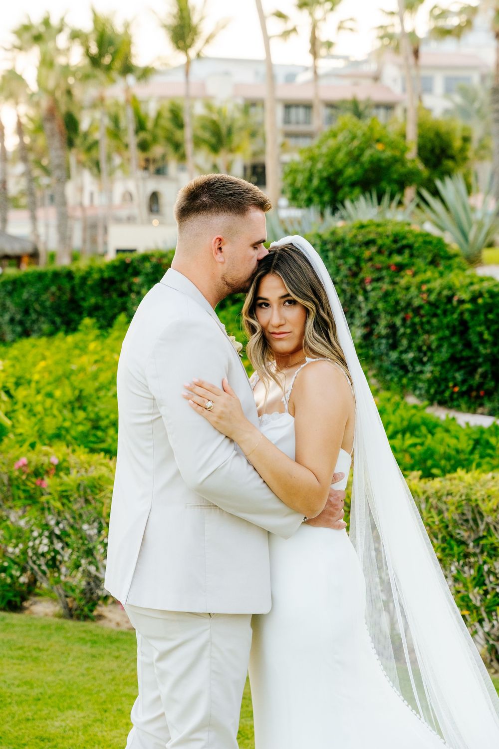 Groom in a light suit kisses the forehead of a bride in a white gown and long veil while embracing in a lush tropical garden.