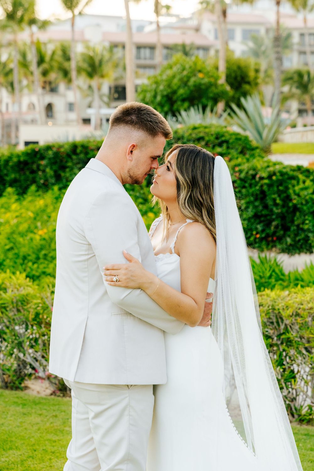 Bride in a white gown and veil embraces her groom in a cream suit, foreheads touching, in a tropical garden with palm trees.