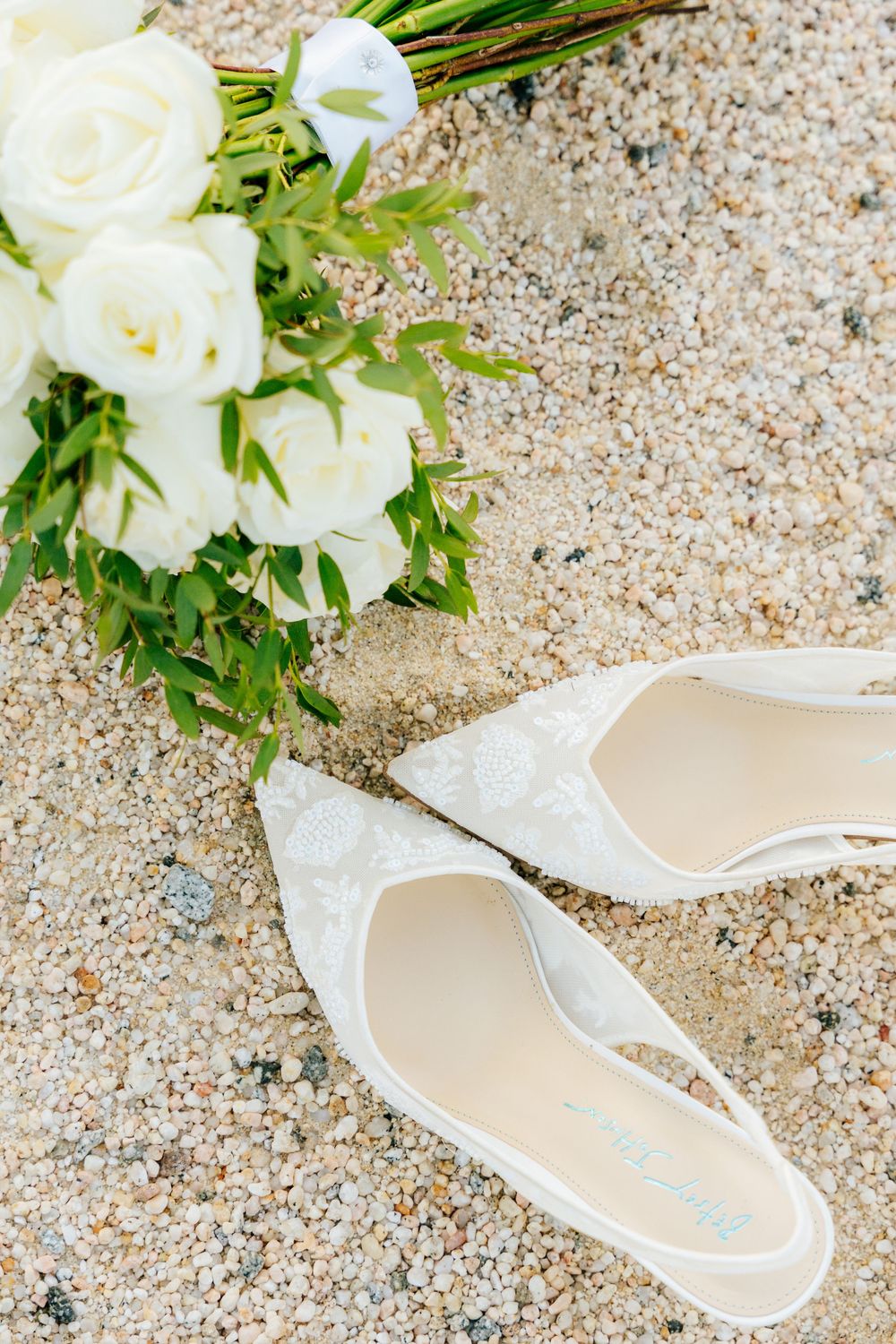White rose bridal bouquet with greenery beside lace pointed-toe wedding heels resting on a pebbly sandy surface.