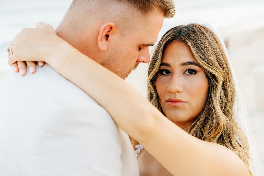 Bride in a long veil wraps her arms around a groom in a light suit during a romantic beachside wedding portrait.