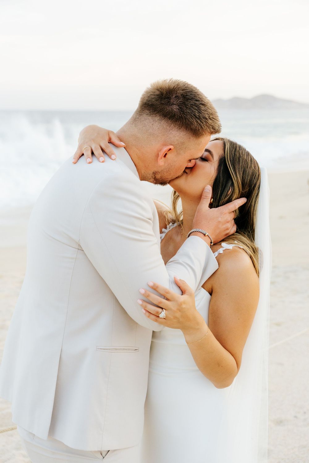 Bride in a white gown and veil kisses groom in a cream suit on a beach as ocean waves roll in at sunset.