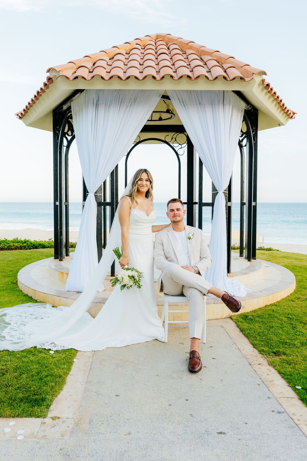 Bride in a white gown holding a bouquet stands beside a seated groom in a beige suit at a beachfront gazebo with ocean views.