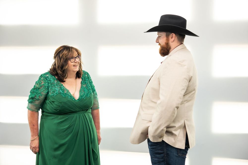 A woman in an emerald green lace dress smiles at a bearded man in a black cowboy hat, cream blazer and jeans.