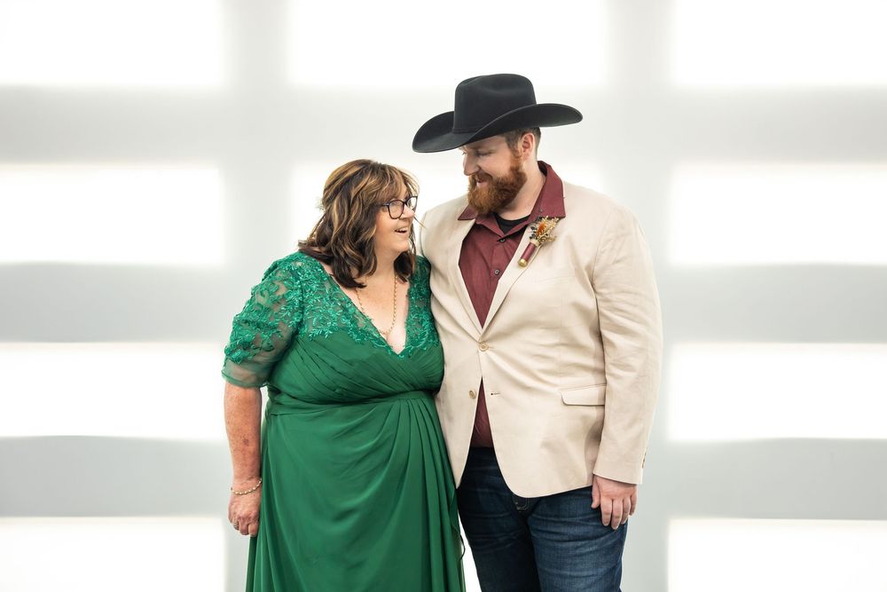 Smiling couple posing close together, a woman in an emerald green lace dress and a bearded man in a cream blazer and black cowboy hat.