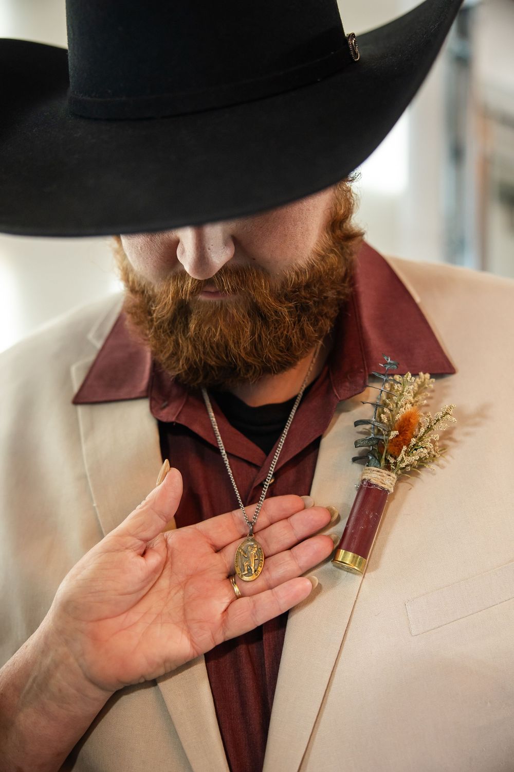 Bearded groom in a black cowboy hat and cream suit jacket holds a pendant necklace, with a shotgun shell boutonniere pinned to his lapel.