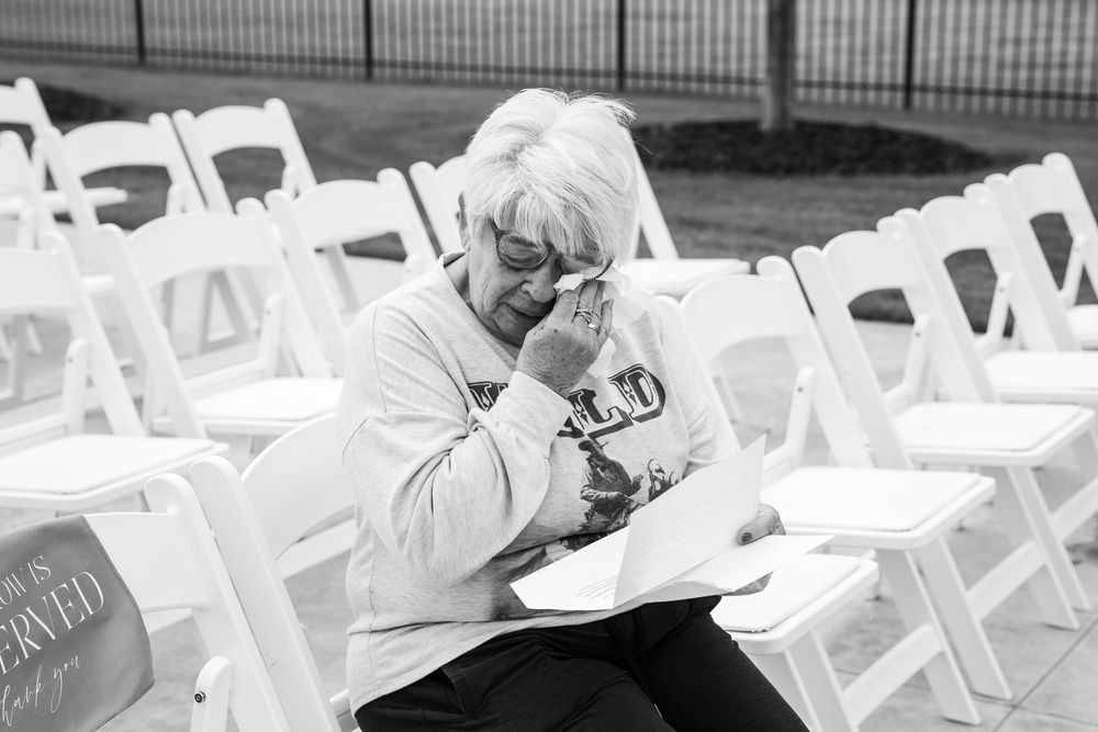 Elderly woman in glasses wipes her eye while holding papers, seated among rows of white folding chairs at an outdoor memorial event.