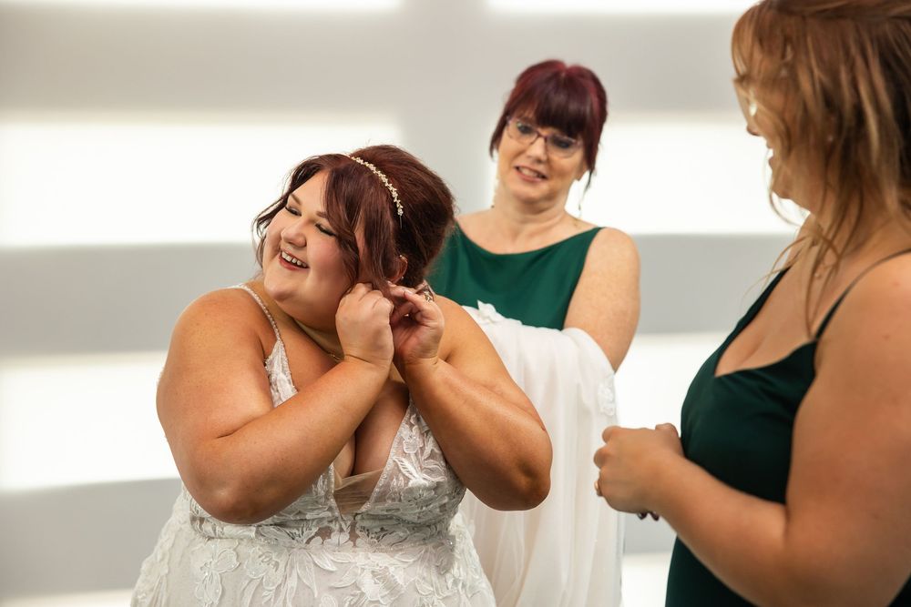 A smiling bride in a white lace gown adjusts her necklace while two bridesmaids in green dresses look on in a bright room.