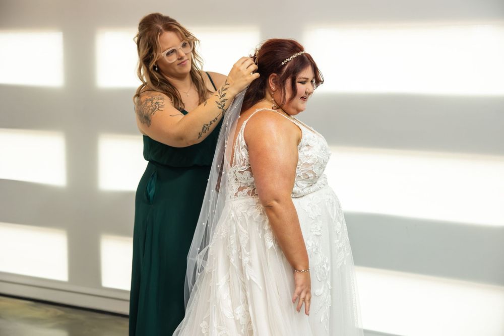 A bridesmaid in a green dress adjusts the veil of a bride wearing a white lace wedding gown during wedding day preparations.