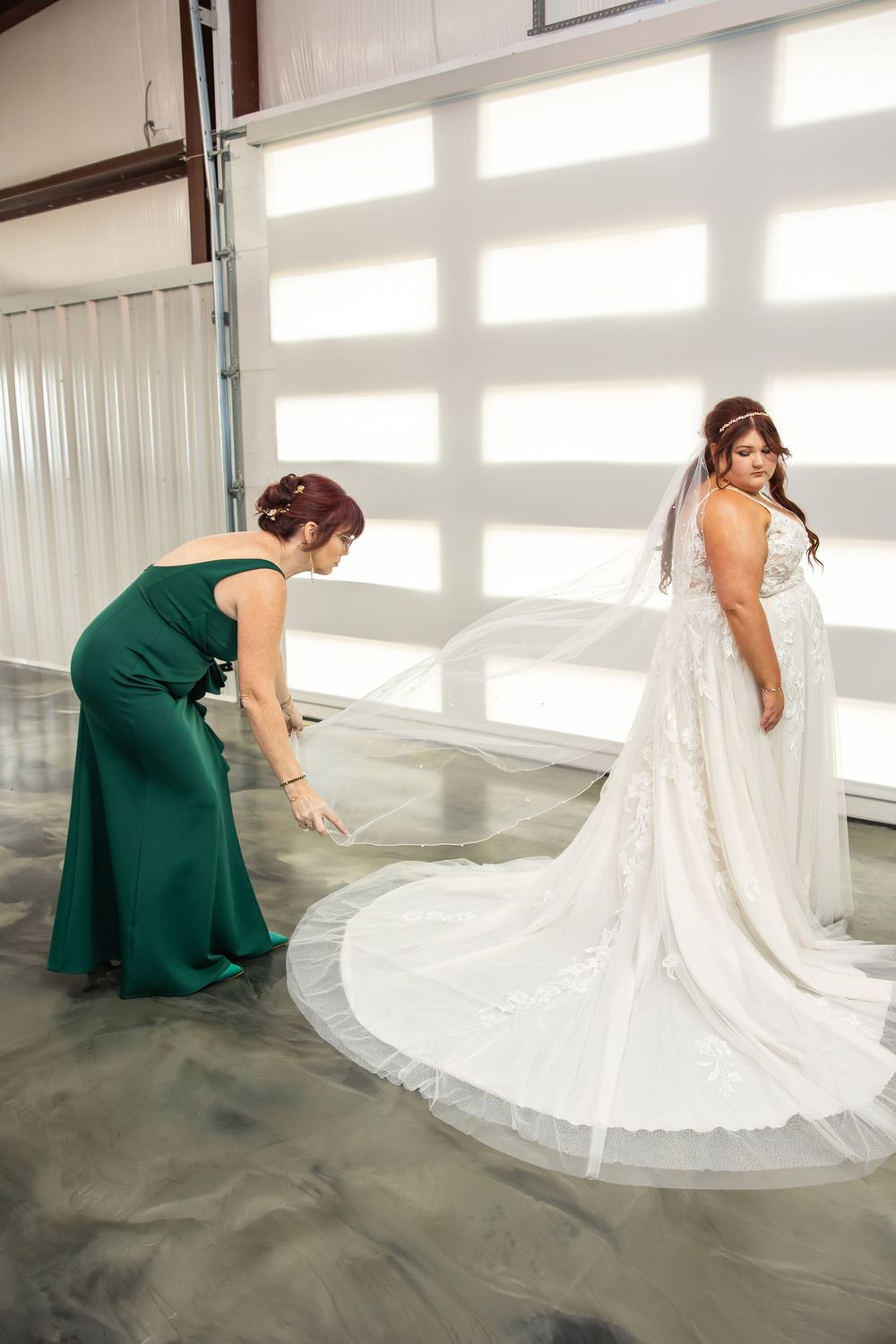Bridesmaid in an emerald green gown adjusts the long cathedral veil and train of a bride in a white lace wedding dress.