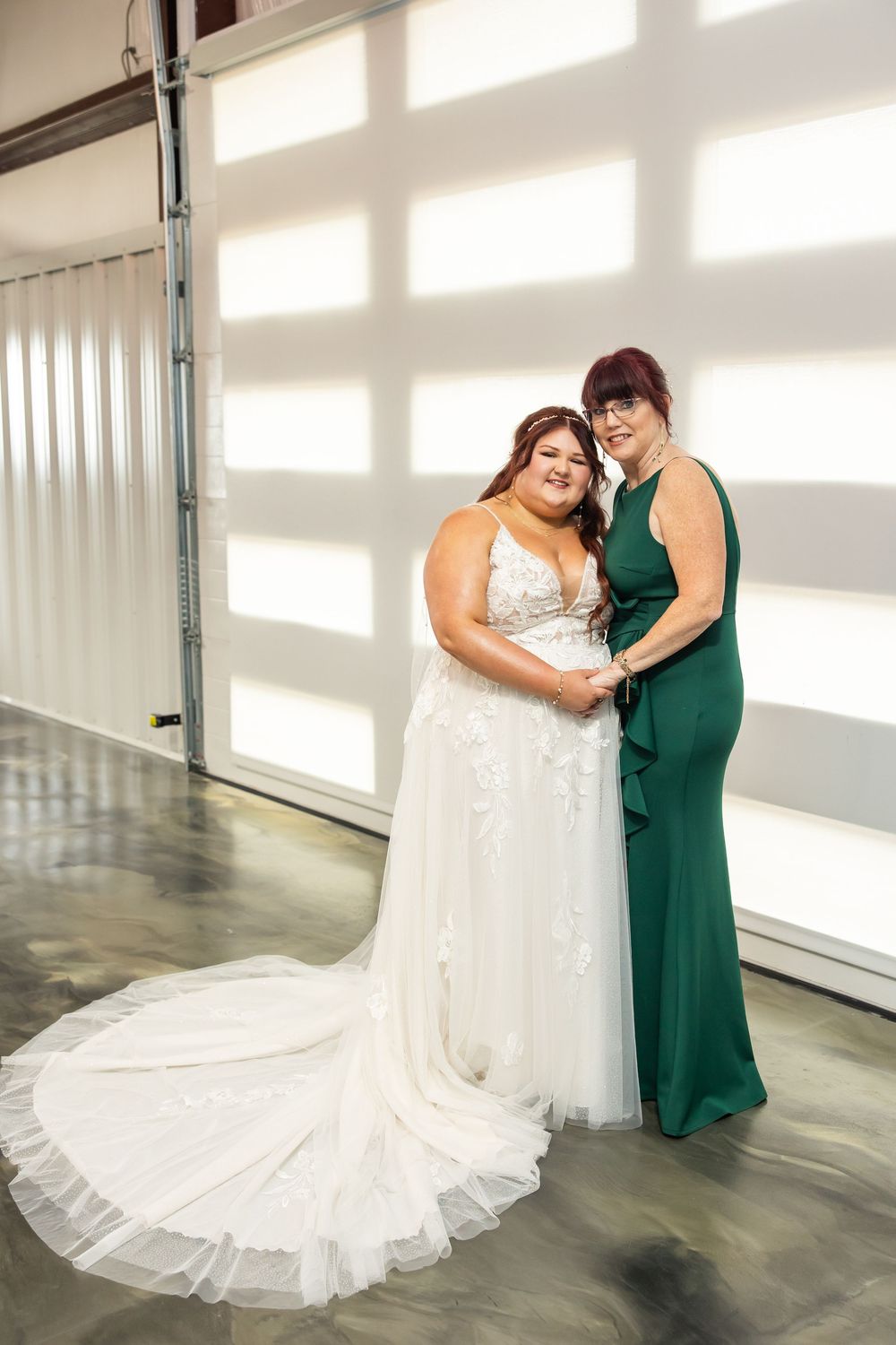Bride in a lace A-line wedding gown with a long train holds hands with a woman in an emerald green dress inside a bright, modern venue.