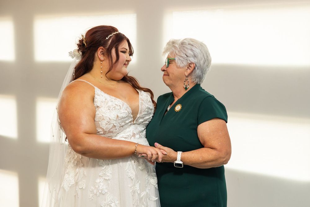 A bride in a white lace wedding gown holds hands with an older woman in a green dress, sharing a tender moment in a bright room.