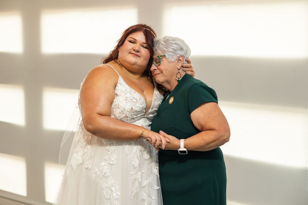 A bride in a white lace wedding gown embraces an older woman in a green dress in an emotional moment against a softly lit wall.