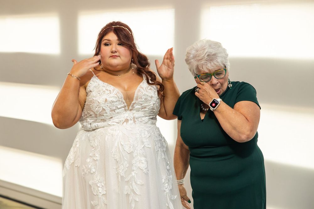 A bride in a white lace gown fans her eyes to hold back happy tears beside an emotional older woman in a green dress and glasses.