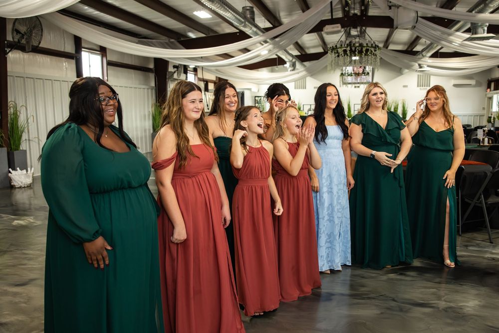 Group of bridesmaids in emerald green and rust red dresses smile and look on with excitement inside a rustic wedding venue with chandeliers.
