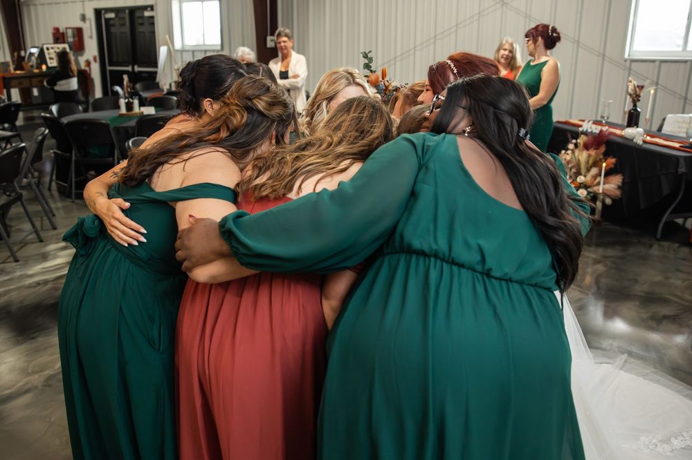Bridesmaids in green and rust dresses stand in a close group hug inside a rustic reception venue, with guests gathered in the background.